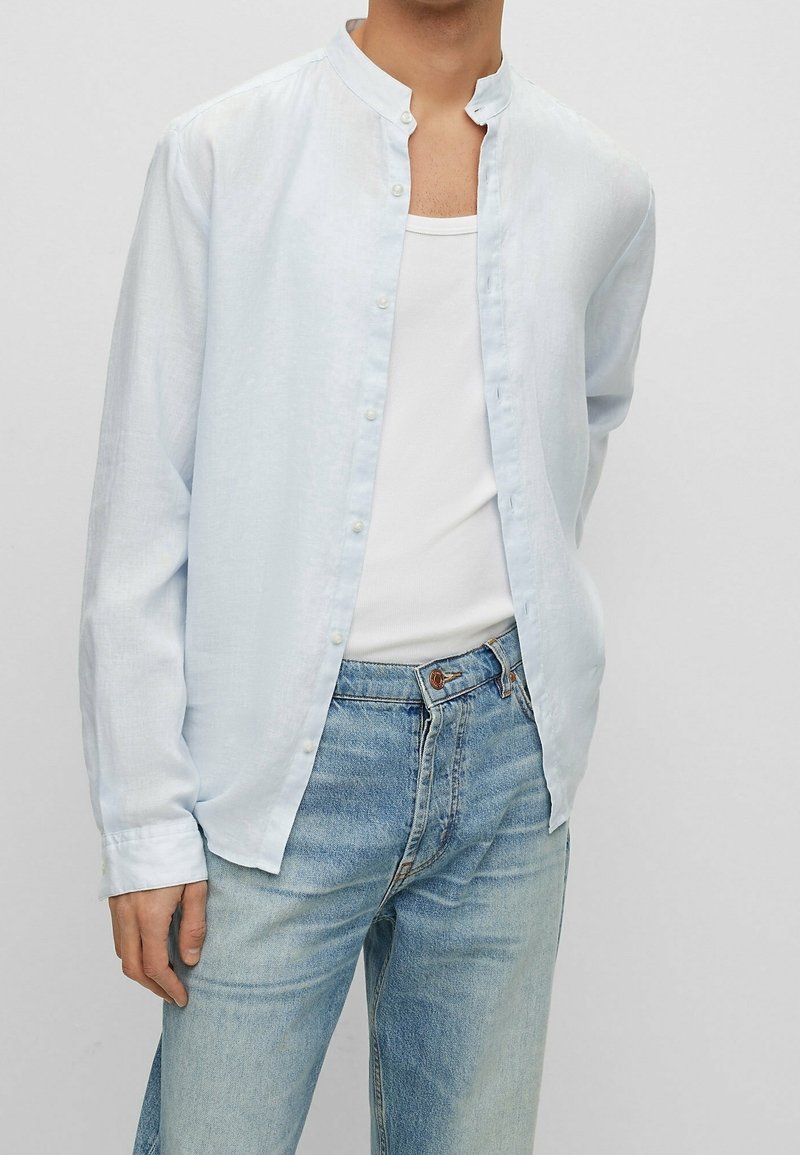 Homme portant une chemise bleu clair à col mandarin sur un débardeur blanc et un jean bleu clair, debout avec un bras derrière le dos.
