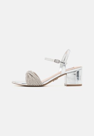 Sandale à talon block argentée avec une bride en perles au niveau des orteils et une fine bride autour de la cheville. Fini métallique brillant.