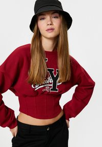Rode crop sweatshirt met zwarte lettering en grafische details, met een gestructureerde ribbel onderkant, gecombineerd met zwarte broek en een zwarte bucket hat.