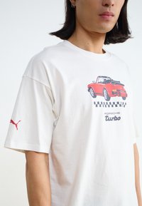 Valge puuvillane T-särk, millel on punane Porsche graafika ja must ruuduline muster. Punane Puma logo vasakul varrukal. Lühikesed varrukad, ümar kaelus.
