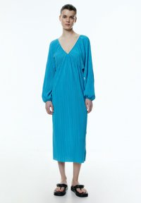 Turquoise jurk met lange mouwen, V-hals, geplooide textuur, midi-lengte en een relaxte pasvorm. Gecombineerd met zwarte platform sandalen.
