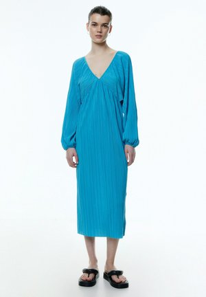 Turquoise jurk met lange mouwen, V-hals, geplooide textuur, midi-lengte en een relaxte pasvorm. Gecombineerd met zwarte platform sandalen.