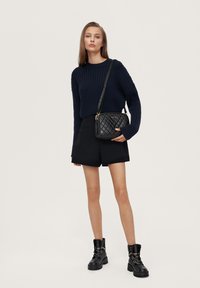 Geantă crossbody neagră matlasată cu accesorii aurii, asortată cu un pulover albastru în dungi și pantaloni scurți negri. Modelul stă în botine negre până la glezne.