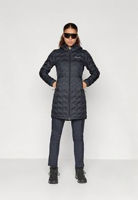 Giacca imbottita nera con cuciture a chevron, zip integrale e collo alto. Indossata con pantaloni blu navy e stivali neri, abbinata a occhiali da sole.