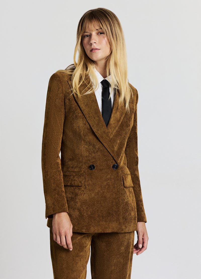 Blazer croisé en velours côtelé marron avec boutons noirs, revers crantés et deux poches avant. Associé à une chemise blanche et une cravate noire.