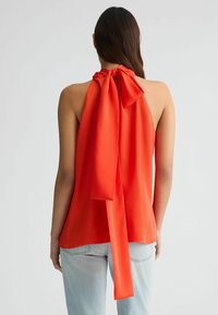 Rotes Satin-Haltertop mit einer gro�ßen Schleifendetail auf der Rückseite. Der Stoff ist glatt und fließend, kombiniert mit hellblauen Jeans.