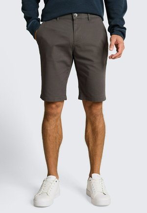 Man draagt donkerbruine knielange shorts, een marineblauwe trui en witte sneakers, staat met zijn handen nabij de zakken tegen een effen achtergrond.