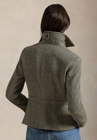 Kurtka w wzór jodełki w kolorach oliwkowym i kremowym, wykonana z tkaniny o splocie, z wysokim kołnierzem i dopasowaną talią. Z tyłu znajduje się szew i ozdobny guzik.