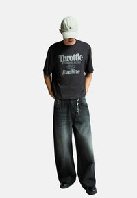 Črna grafična majica s napisom "Throttle Redline Rush", kombinirana s širokimi, zbledelimi črnimi kavbojkami in črnimi čevlji, na vrhu pa lahka kapa v svetli barvi.