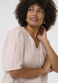 Blusa rosa chiaro con maniche a sbuffo, scollo a V e quattro piccoli bottoni. Tessuto morbido e testurizzato aggiunge un dettaglio sottile.