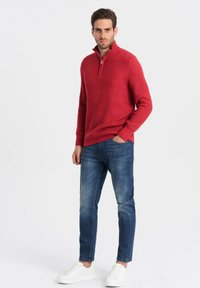 Rode gestructureerde pullover met een halflange ritssluiting, gecombineerd met blauwe slim-fit jeans en witte sneakers, tegen een effen achtergrond.