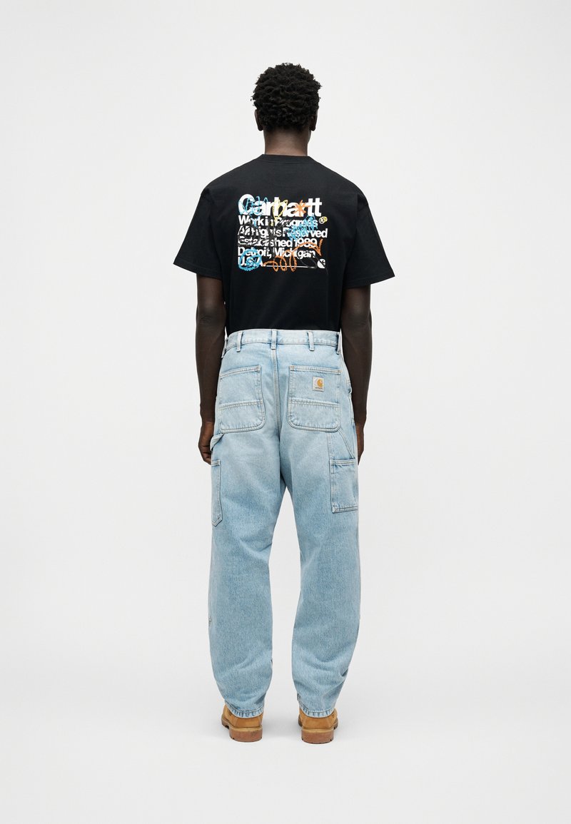 Personne vue de dos portant un t-shirt graphique noir avec un texte coloré, un jean large bleu clair avec des poches, assorti à des bottes de travail marron clair.