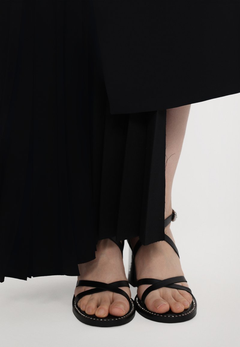 Gonna nera a pieghe e sandali piatti. I sandali presentano cinturini incrociati e un bordo decorativo con borchie attorno alla suola.