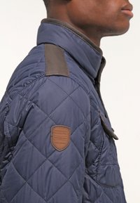 Marineblå quiltet jakke med brun læderlap på skulderen og detaljer. Stoffet har en glat tekstur med en struktureret krave.