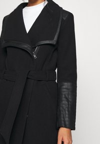 Abrigo negro con detalles de cuero sintético en el cuello y las mangas, cintura con cinturón, parches texturizados en los codos y detalle de cremallera en la parte delantera.