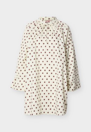 Kremowa bluzka z długim rękawem w brązowe kropki, z wysokim kołnierzem z ozdobą w postaci kokardy oraz mankietami zapinanymi na guziki na jasnoszarym tle.
