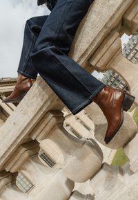 Stivali alla caviglia in pelle marrone con tacco a blocco. Indossati con pantaloni di jeans scuri, appoggiati su un parapetto di pietra in un ambiente all'aperto.