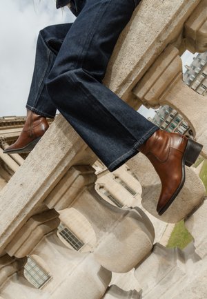 Braune Leder-Stiefeletten mit Blockabsatz. Getragen mit einer dunklen Jeans, die auf einem Steinbalkon in einer Outdoor-Umgebung ruhen.