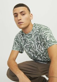 Groen T-shirt met korte mouwen en een wit loofpatroon. De stof lijkt lichtgewicht met een gladde textuur. Het model zit in een hurkende positie en draagt een paarse broek.