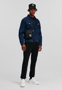 Blauwe denimjacket, zwarte bucket hat, zwarte crossbody tas, zwarte broek en witte sneakers. Minimalistisch ontwerp met merkaccenten.