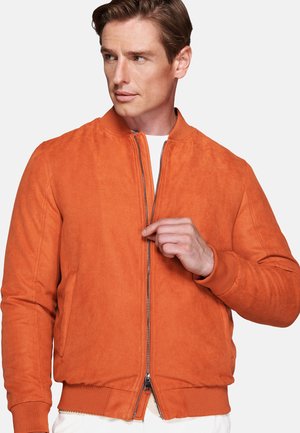 Man wearing an orange suede bomber jacket partially zipped over a white shirt, looking to his left.