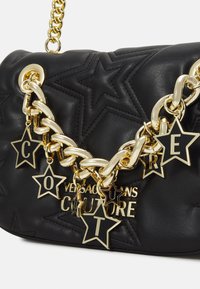 Sac à main en cuir noir avec un motif matelassé en étoiles, bandoulière en chaîne dorée et pendentifs décoratifs en forme d'étoile affichant des lettres. Présente un logo distinctif.