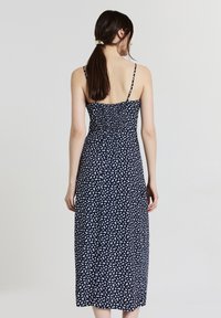 Femme aux longs cheveux bruns attachés en queue de cheval, portant une robe midi bleu marine avec un motif floral blanc, de dos contre un fond léger uni.