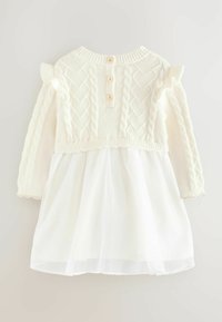 Krämfärgad klänning i stickad stil med texturerad bodice, långa ärmar, volangdetaljer och en genomskinlig vit kjol. Har tre knappar i trä på baksidan.