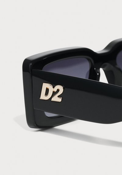 Sorte solbriller med en tyk ramme, der har mørke linser og et guld "D2" logo på siden. Glat, blank overflade og dristigt design.