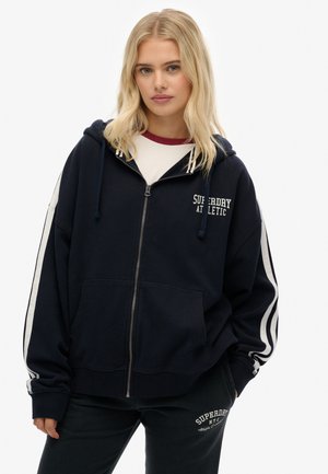 Blonde vrouw in een marineblauwe hoodie met rits en witte strepen op de mouwen, en bijpassende marineblauwe joggingbroek met witte tekstlogo's.