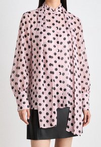 Blouse rose clair en tissu transparent avec des pois noirs, manches longues, fermeture à boutons et détail de nœud drapé au cou.