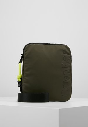 Kwadratowa oliwkowozielona saszetka Calvin Klein z czarnym zamkiem błyskawicznym, neonowo-żółtym uchwytem zamka i czarnym paskiem na nadgarstek na białej powierzchni.