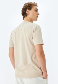 T-shirt di cotone beige con maniche corte, collo girocollo e una texture liscia. Cuciture visibili sull'orlo e sulle maniche, indossata da un modello.