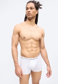 Μυώδης άνδρας με dreadlocks που φοράει λευκά εσώρουχα Tommy Hilfiger, στέκεται με τα χέρια χαλαρά έναντι ενός απλού φωτεινού φόντου.