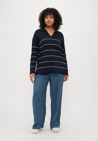 Marineblauw long-sleeve shirt met witte horizontale strepen, voorzien van een V-hals. Losvallende lichtblauwe broek en witte schoenen maken de outfit compleet.