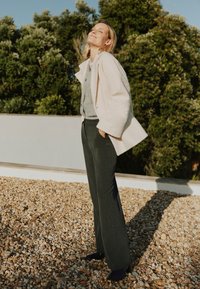 Abrigo texturizado de color beige claro sobre un suéter gris, combinado con pantalones gris oscuro con remaches laterales, y zapatos de tacón negros. Ambiente exterior.