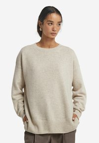 Beiger, oversize Strickpullover mit rundem Ausschnitt, gerippten Bündchen und Saum. Weiche Textur und gestuftes Design. Getragen über dunklen Hosen.