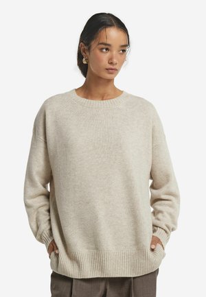 Beżowy, oversize sweter dzianinowy z okrągłym dekoltem, ściągaczami przy mankietach i u dołu. Miękka tekstura i warstwowy design. Nosi się go na ciemnych spodniach.