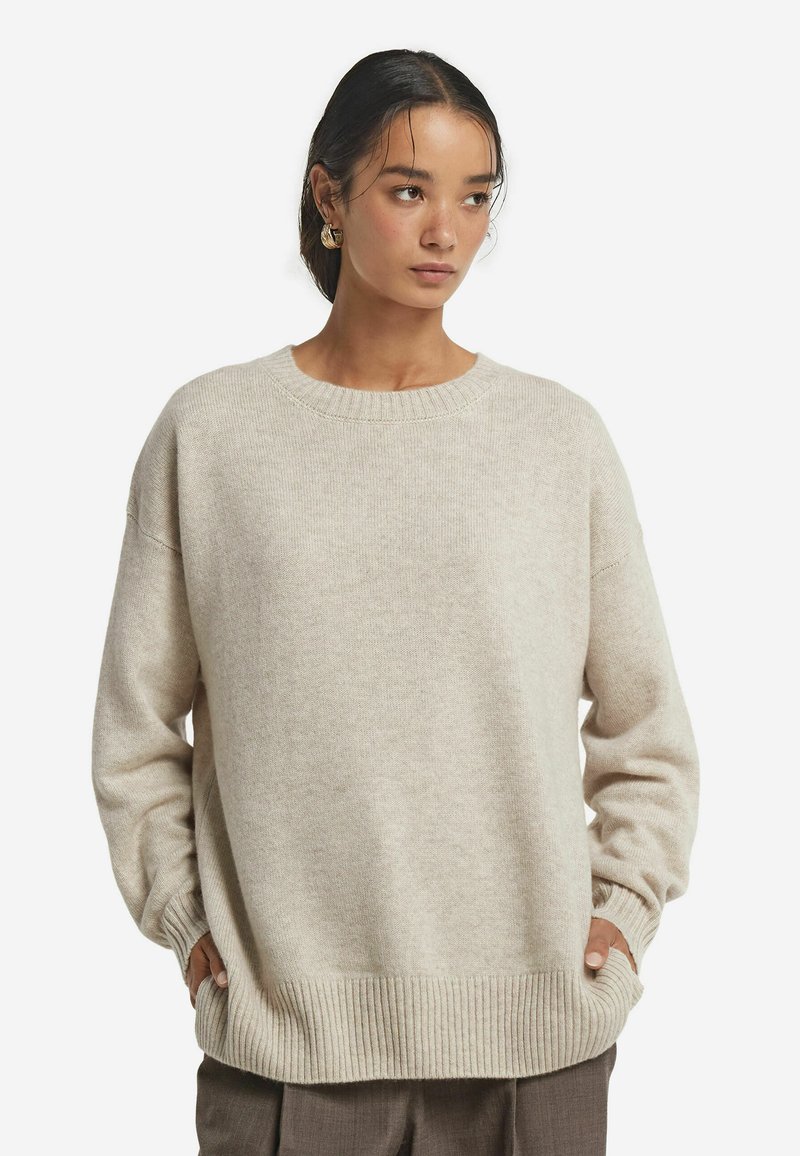 Beiger, oversize Strickpullover mit rundem Ausschnitt, gerippten Bündchen und Saum. Weiche Textur und gestuftes Design. Getragen über dunklen Hosen.