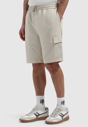 Man draagt beige cargobroek met trekkoord in de taille, witte sokken met zwarte letters en witte sneakers met rubberen zolen.