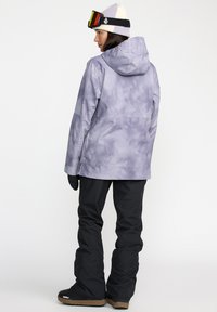 Chaqueta de snowboard con diseño tie-dye púrpura y capucha, combinada con pantalones de nieve negros. Botas negras con una suela marrón visible en la parte inferior.