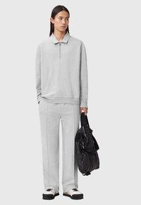 Helles graues Sweatshirt mit einem Reißverschlusskragen, kombiniert mit passender weit geschnittener Sweatpants. Schwarzer und grauer Rucksack mit Trägern. Klobige weiße Sneakers.