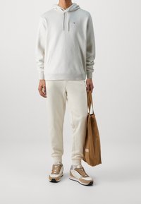 Vit hoodie med huva och ficka fram, tillsammans med ljust beige sweatpants och bruna sneakers. Håller en brun tygväska.