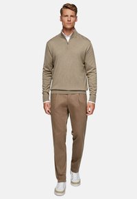 Maglione beige con zip e texture a coste, abbinato a pantaloni marrone chiaro e sneakers bianche. Modello in piedi su uno sfondo neutro.