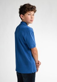 Modrá polo tričko s krátkým rukávem, vyrobené z bavlny; má klasický límec a boční rozparky na spodním lemu. Nosit s tmavými džíny.