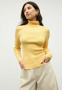 Frau in gelbem, geripptem Rollkragensweater und beigefarbenen Hosen, die mit den Händen in den Taschen steht und leicht zur Seite blickt, vor einem schlichten Hintergrund.