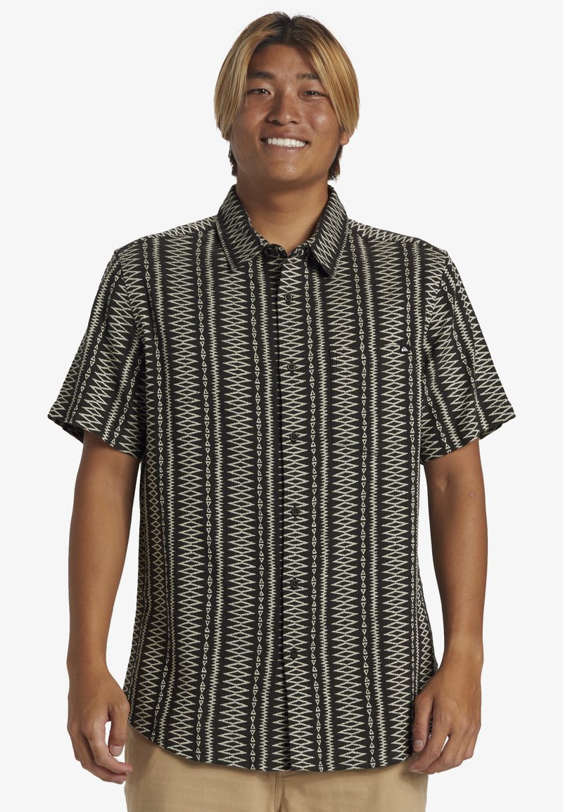Camisa de manga corta en negro y crema con un patrón geométrico, que cuenta con un cierre de botones en el frente y un cuello clásico. Material de algodón.