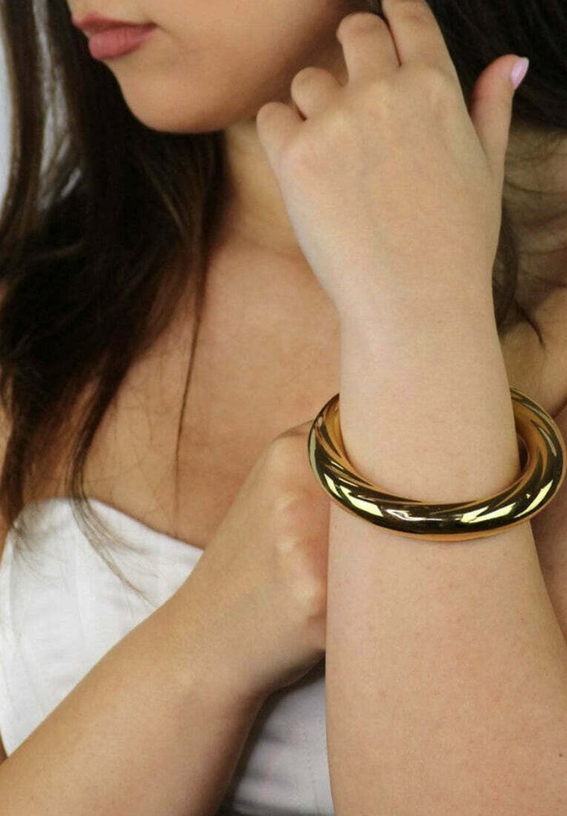 Bracciale in oro con una finitura liscia e lucida, dal design intrecciato, indossato su un polso con pelle leggermente texturizzata.