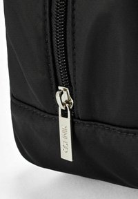 Czarna torba z nylonu z gładkim zamkiem błyskawicznym i srebrnym uchwytem oraz brandingiem, o teksturowanej powierzchni. Struktura jest prostokątna.