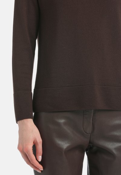 Brauner Strickpullover mit langen Ärmeln, der einen strukturierten Stoff und einen gerippten Saum aufweist. Kombiniert mit glänzenden, dunkelbraunen Lederhosen.